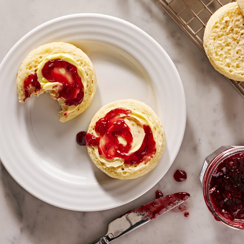 crumpets topped with butter and jam on a white plate beside jam jar and jam knife and rack with more crumpets