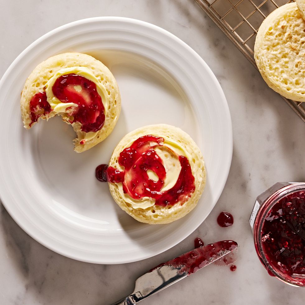 crumpets topped with butter and jam on a white plate beside jam jar and jam knife and rack with more crumpets
