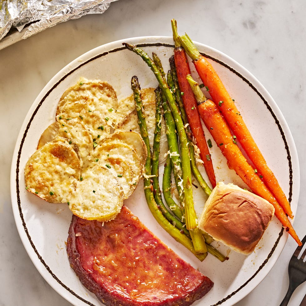 sheet pan easter dinner with glazed ham, gruyere scalloped potatoes, roasted carrots, roasted asparagus, and a roll on a plate