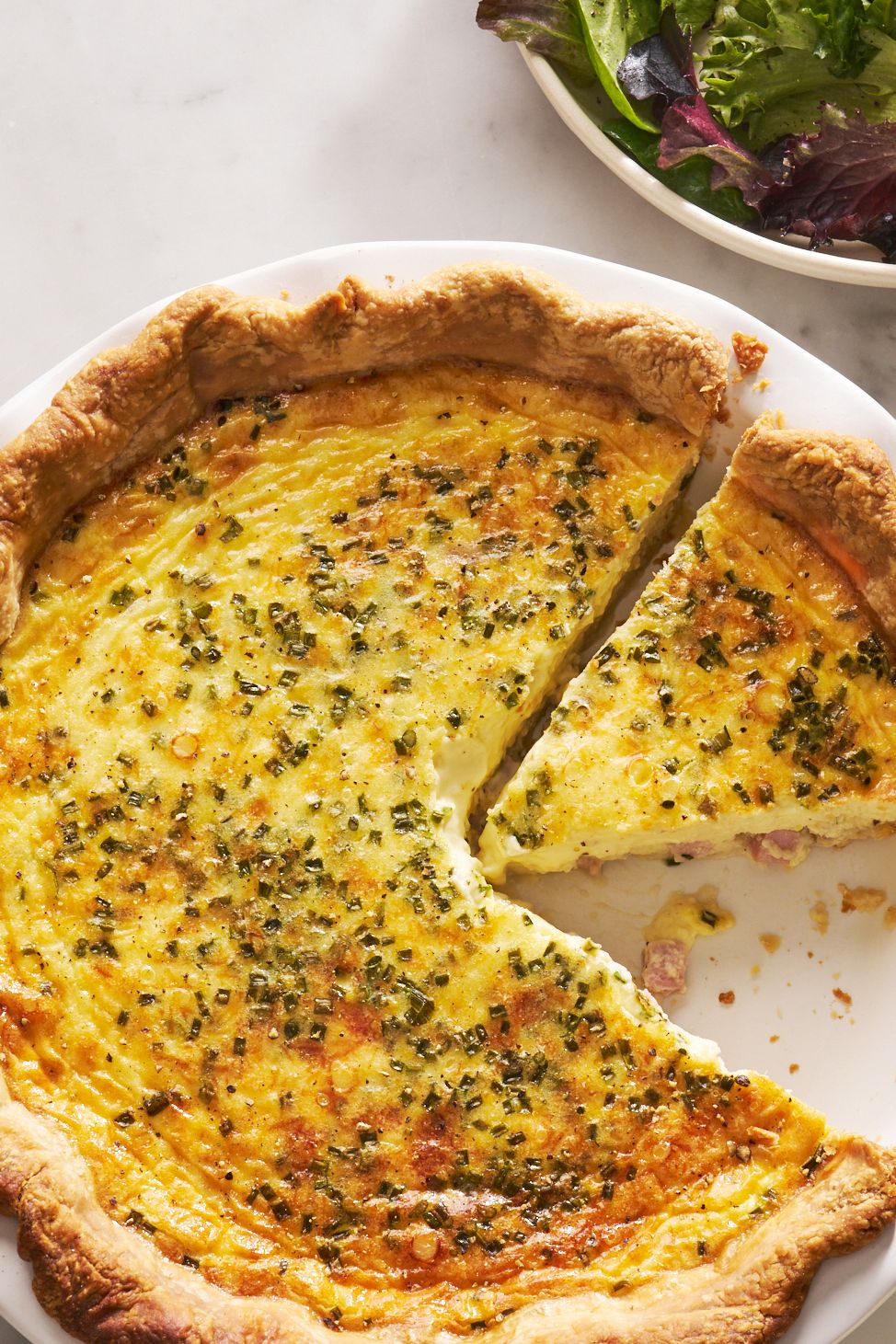 ham and cheese quiche with a slice cut out beside a salad bowl