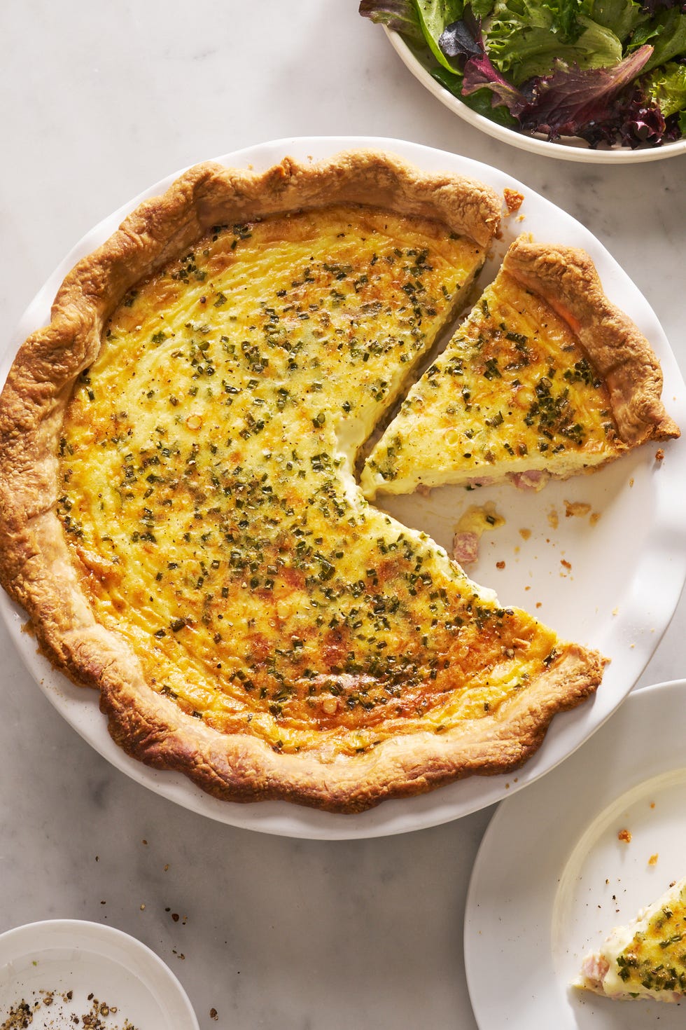 ham and cheese quiche with a slice cut out beside a salad bowl