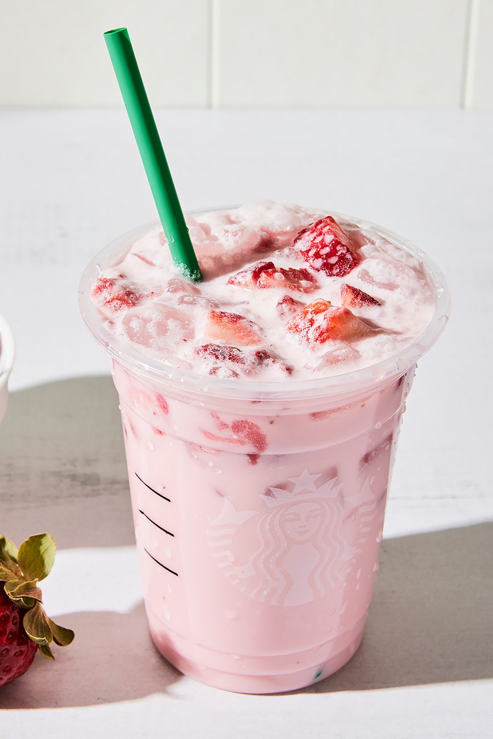 a tall glass with creamy light pink beverage on ice with sliced fresh strawberries and a glass straw