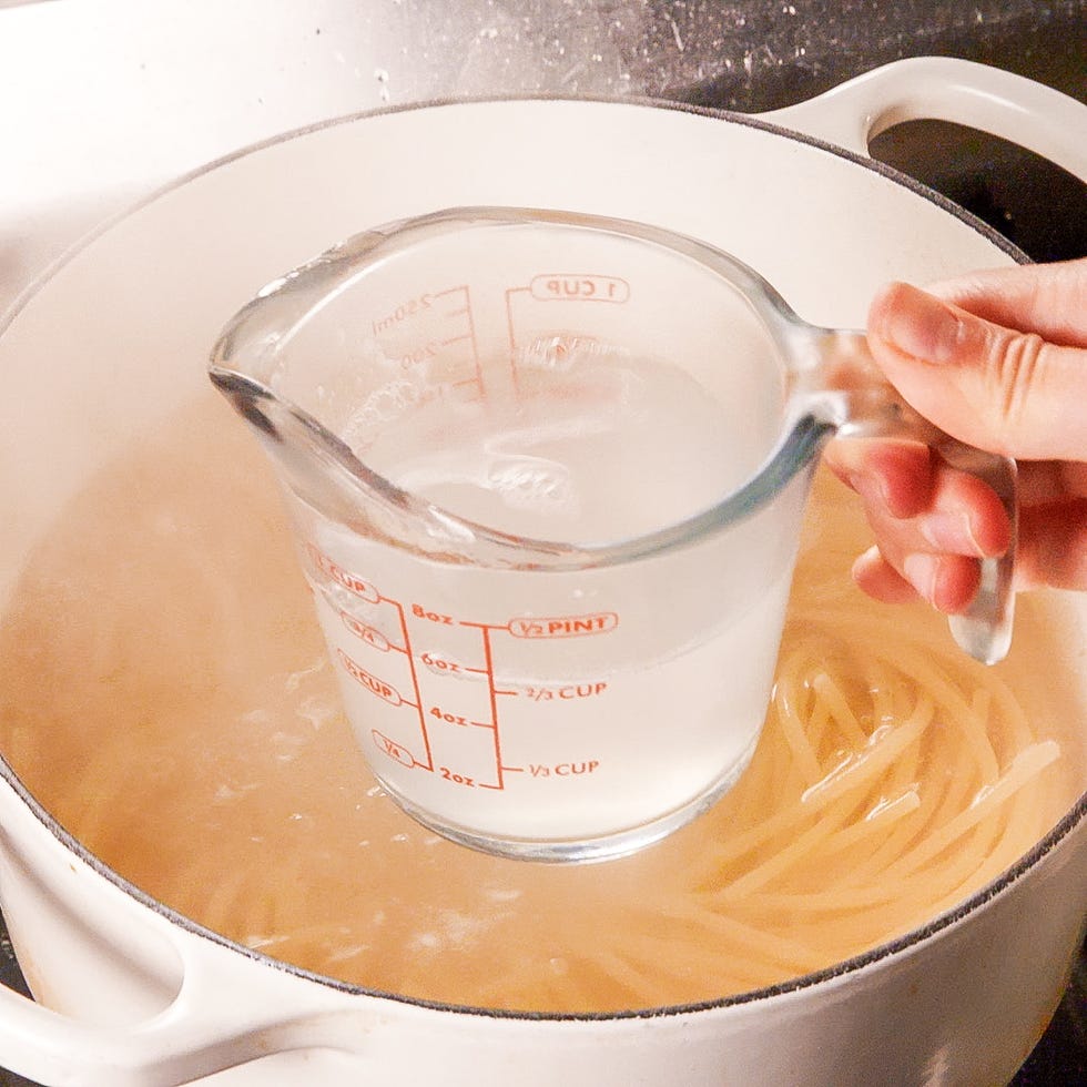 hand holding a measuring cup with water above a pot of simmering pasta