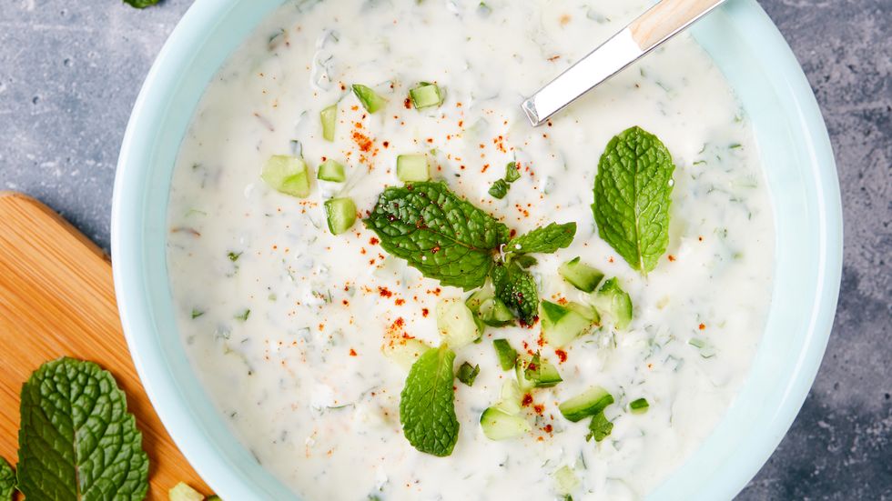 raita in a bowl with cucumbers, mint, and powdered red spice cutting board with mint and cucumbers alongside