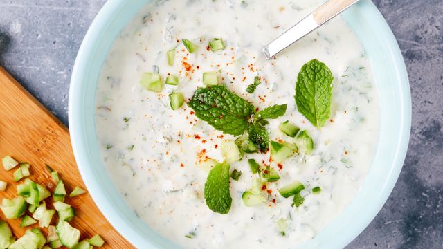 raita in a bowl with cucumbers, mint, and powdered red spice cutting board with mint and cucumbers alongside