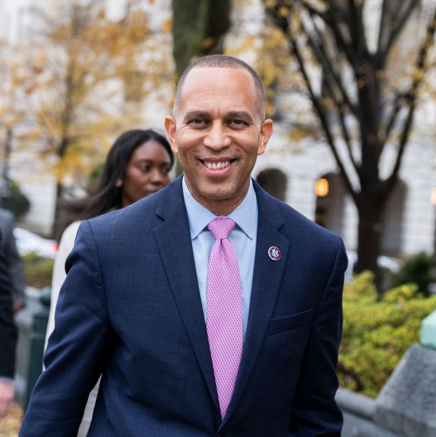 hakeem jeffries smiles at the camera as he walks on a city sidewalk, he wears a navy suit jacket with a light blue collared shirt and a pink tie