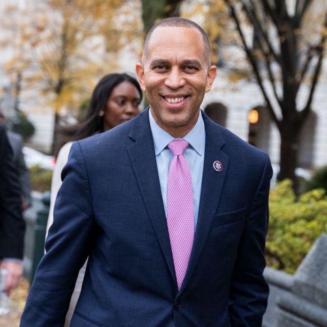 hakeem jeffries smiles at the camera as he walks on a city sidewalk, he wears a navy suit jacket with a light blue collared shirt and a pink tie