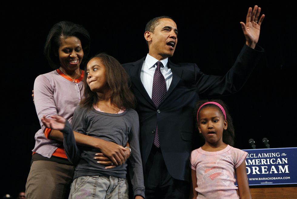 standing on a stage in front of a podium, michelle embraces malia obama from behind while barack obama waves his hand in the air above sasha obama
