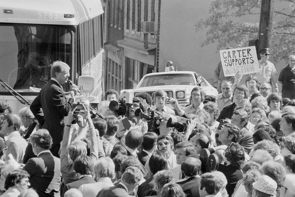 jimmy carter stands among a crowd and speaks into a microphone, behind him is a bus and police car