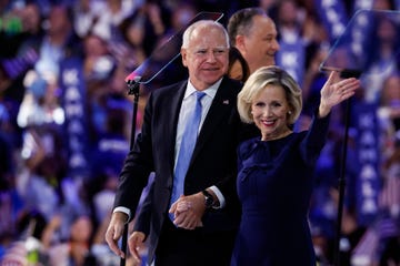 tim and gwen walz stand on a stage as they smile and hold hands, she waves