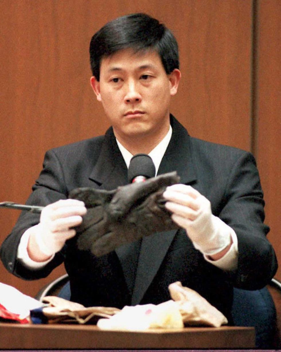dennis fung sits on the witness stand and holds a black glove with his hands in white gloves, he wears a dark suit jacket and white collared shirt