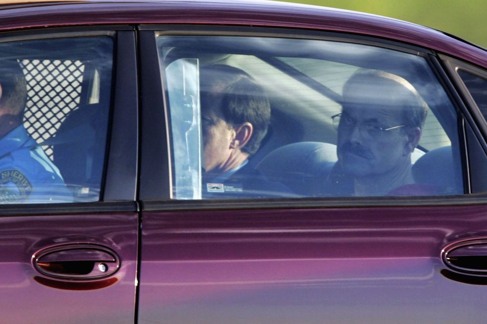 dennis rader looks out the backseat window of a purple car, he wears glasses and two other men are partially visible elsewhere in the car