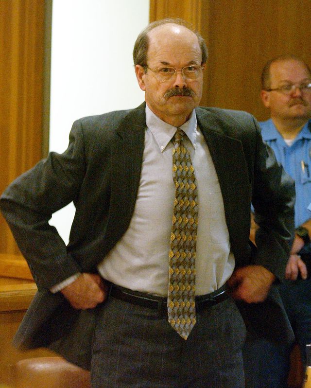 btk killer dennis rader tugs at his waistband while standing in a wood paneled room, he wears a gray suit with a collared shirt and patterned yellow and brown tie, he has a mustache and large glasses