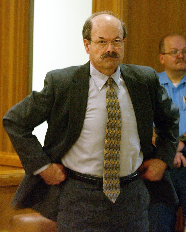 btk killer dennis rader tugs at his waistband while standing in a wood paneled room, he wears a gray suit with a collared shirt and patterned yellow and brown tie, he has a mustache and large glasses