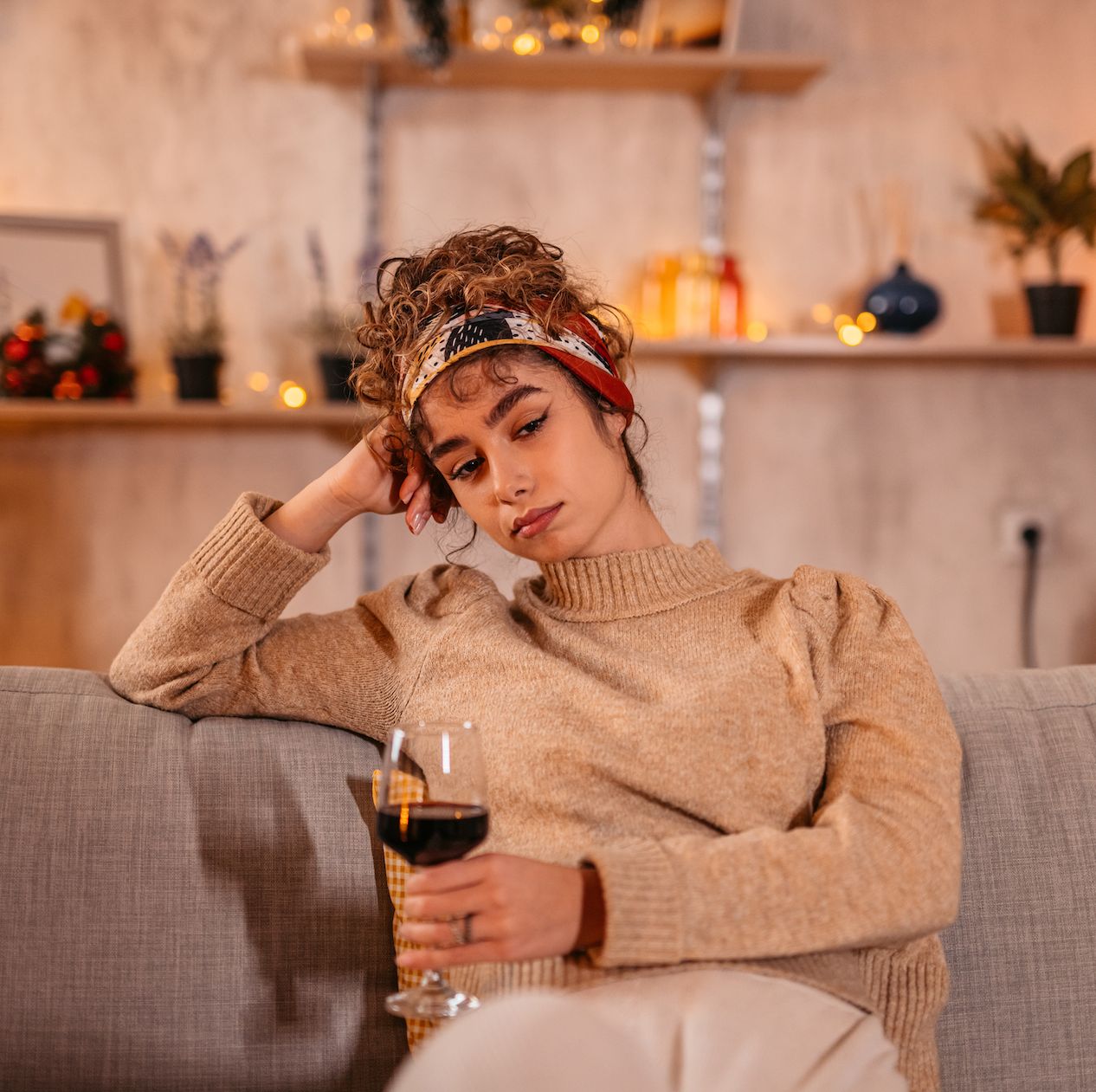 lonely and sad young woman drinking a glass of red wine alone on the sofa with christmas lights and a tree behind her