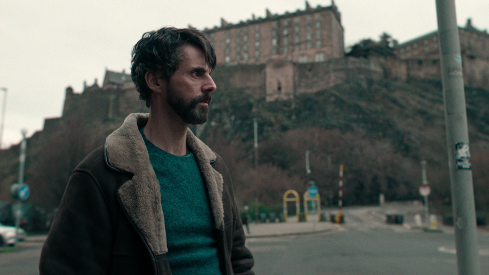 Matthew Goode trägt eine Lammfelljacke mit einem grünen Pullover und steht vor Edinburgh Castle