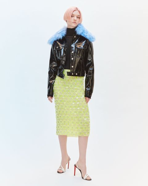 a white woman with short pink hair wearing a shiny black jacket with blue furry collar and chartreuse embroidered skirt