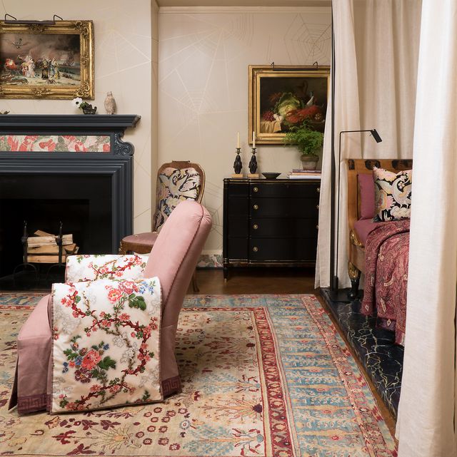 cozy bedroom with a chaise enfolded by tall draperies and a border carpet on wood floors and a pink chair with skirt and what looks like embroidered arm sleeves and a blue mantel fireplace in the back with oil painting on the wall