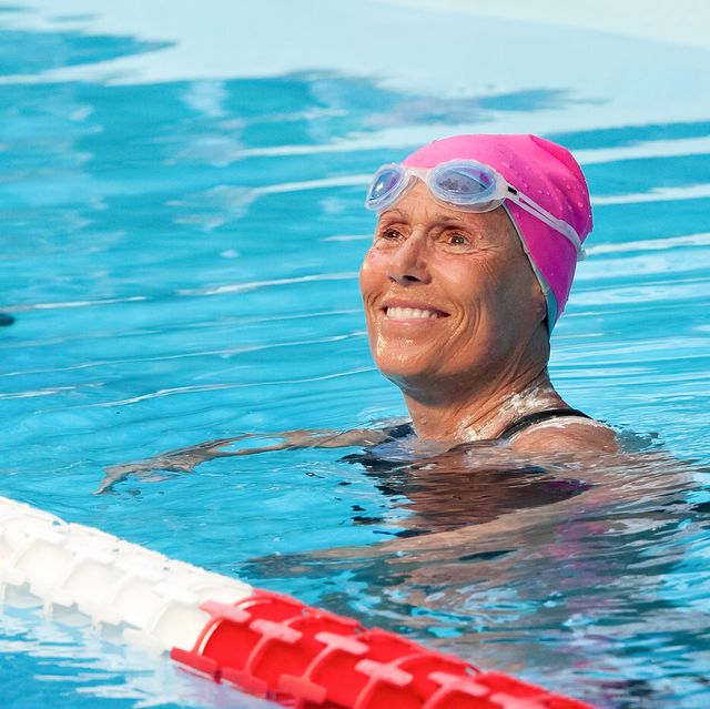diana nyad floats in a swimming pool and looks past the camera smiling, she wears a pink swimming cap and white and blue googles on her forehead