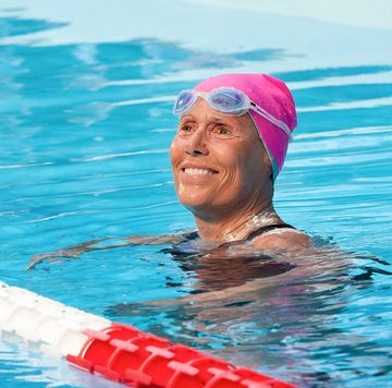 diana nyad floats in a swimming pool and looks past the camera smiling, she wears a pink swimming cap and white and blue googles on her forehead