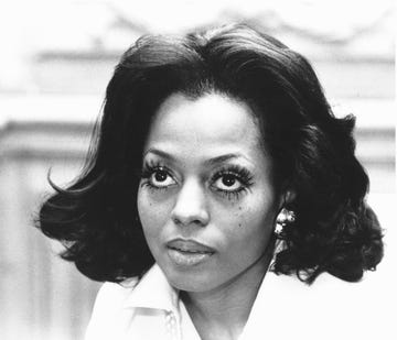 diana ross looks upward with a neutral expression on her face, she has large eyelashes on with a shiny earring peeking from behind her shoulder length hair