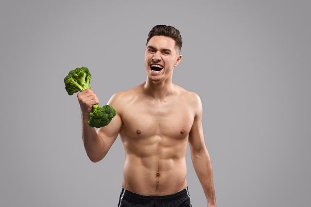 powerful shirtless athlete screaming and looking at camera while exercising with broccoli dumbbell against gray background