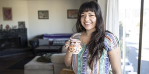 Young Latina Stands Alone in Her Living Room, Wearing Woven Ethnic Blouse, Smiling, Holding a Floral Pottery Mug