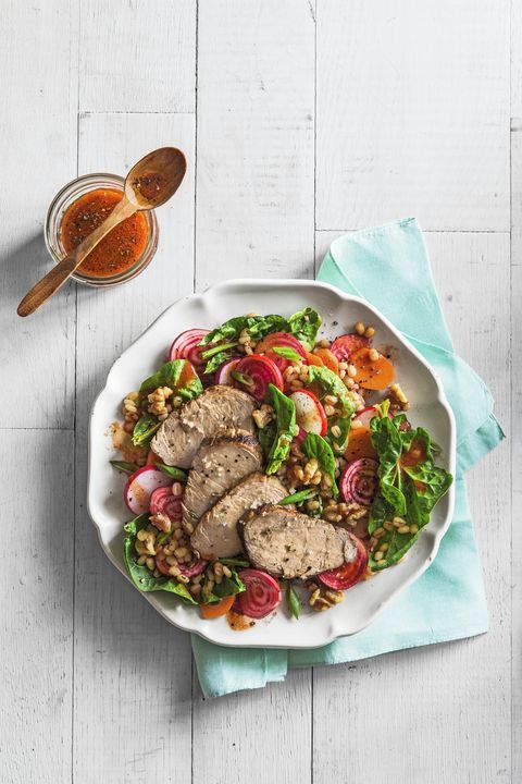 spinach and barley salad with grilled pork in a white bowl on a white wooden surface with a light teal napkin