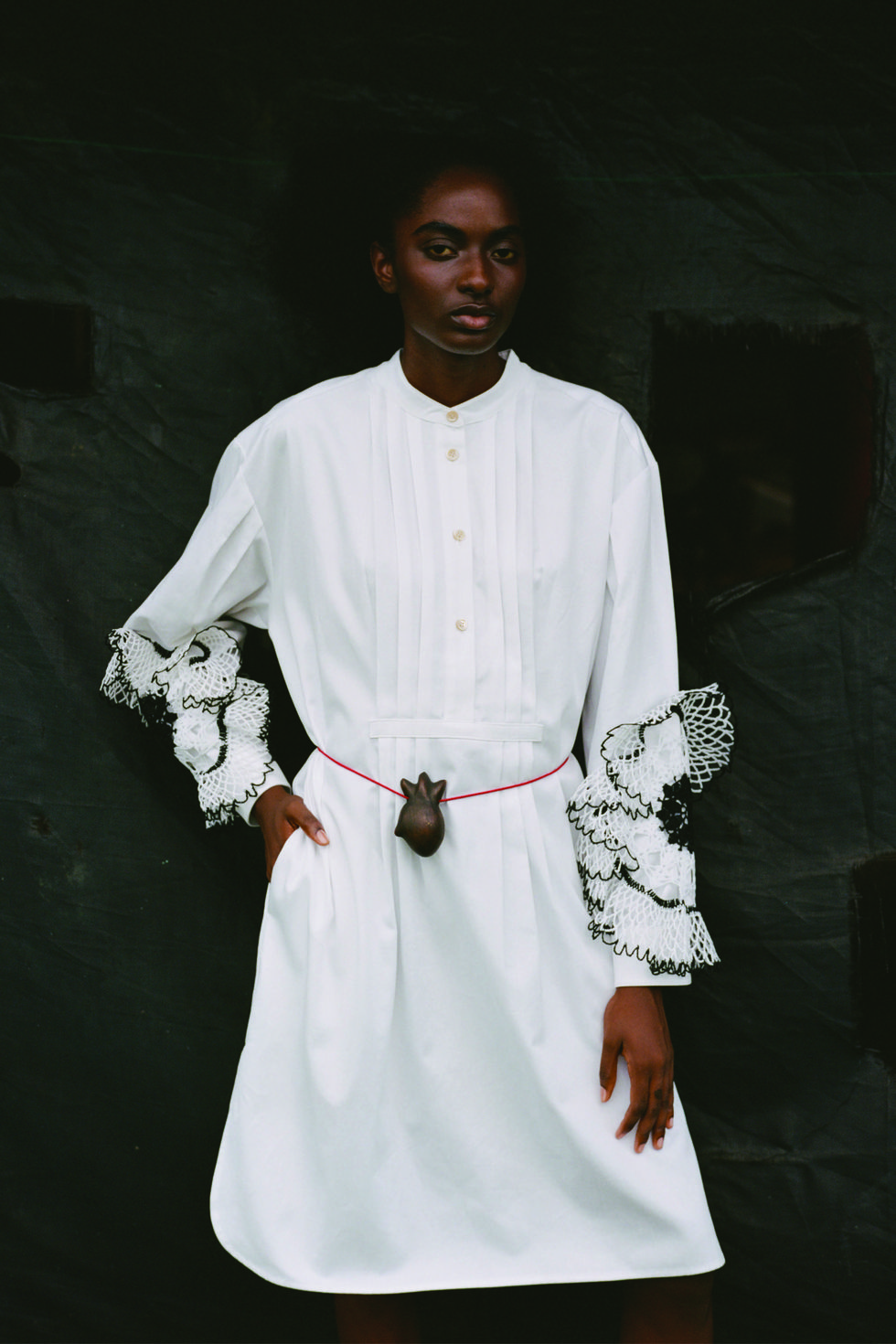 a model in a white shirt dress with a wooden carved human heart worn around her waist poses in front of a black background
