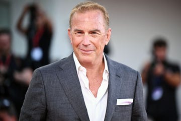 kevin costner smiles at the camera, he wears a gray suit jacket with a white pocket square and a white collared shirt that is partially unbuttoned