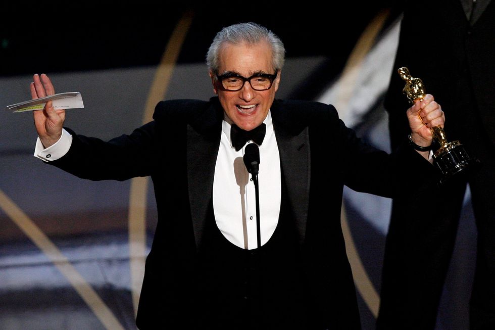 martin scorsese stands on a stage and speaks into a microphone, both his hands are raised and he holds notecards and an oscar statuette, he wears a black tuxedo and bowtie with a white shirt and black glasses