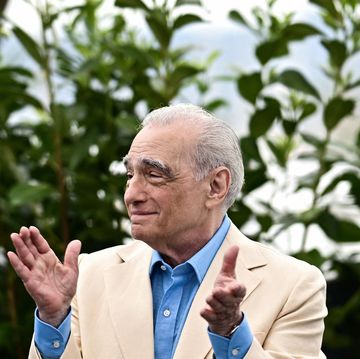 martin scorsese claps his hands while standing in front of greenery, he looks to the left and wears a tan suit jacket over a blue collared shirt