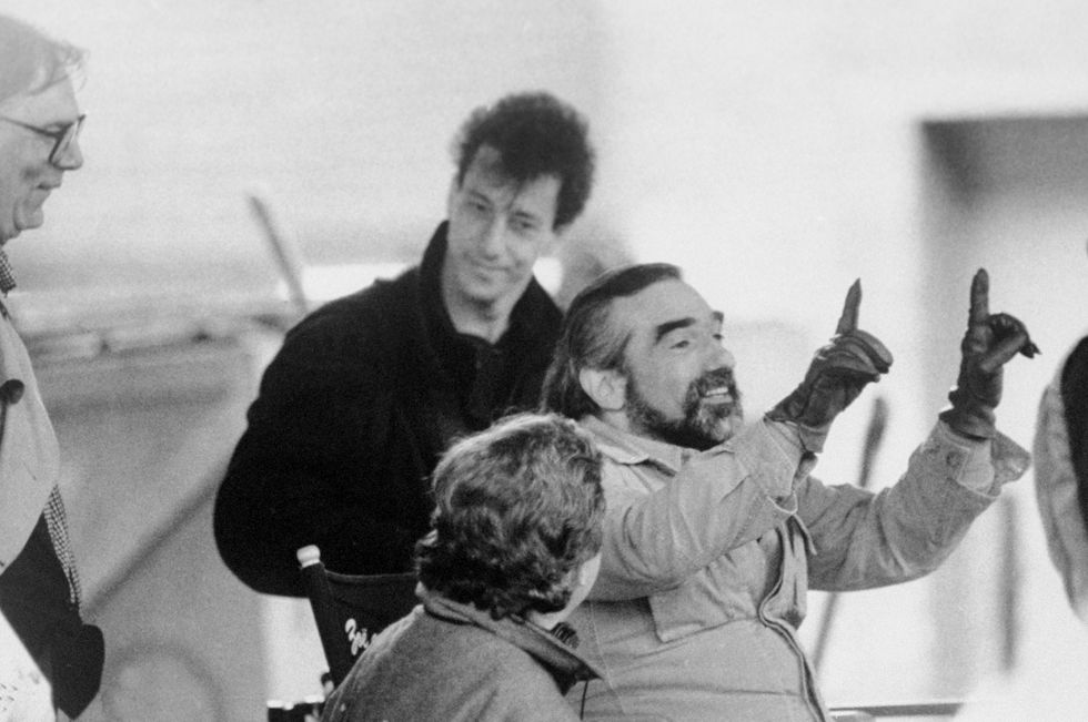 a black and white photo of a bearded martin scorsese pointing upwards and smiling as he directs a film, with several people around him