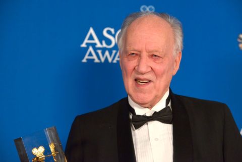 werner herzog, wearing a black tuxedo, stands in front of a blue backdrop and talks