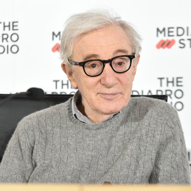 woody allen stars at the camera with a small smile, he wears his signature black framed glasses and a gray sweater over a collared shirt, he is shitting in a chair during a press interview, a microphone enters the frame from the left and behind him is a white background with a repeating logo