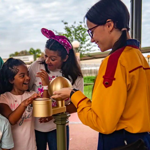 a smiling girl tapping her magicband plus wristband to a kiosk at a walt disney world theme park entrance, with her mother and a cast member smiling back at her