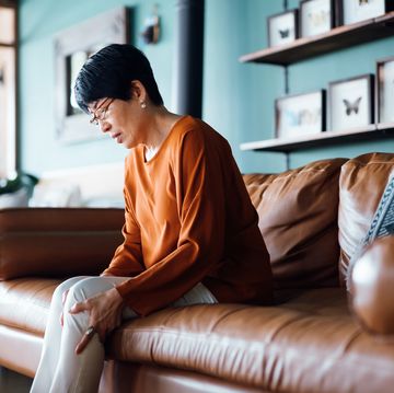 a distraught senior asian woman feeling unwell, suffering from pain in leg while sitting on sofa in the living room at home