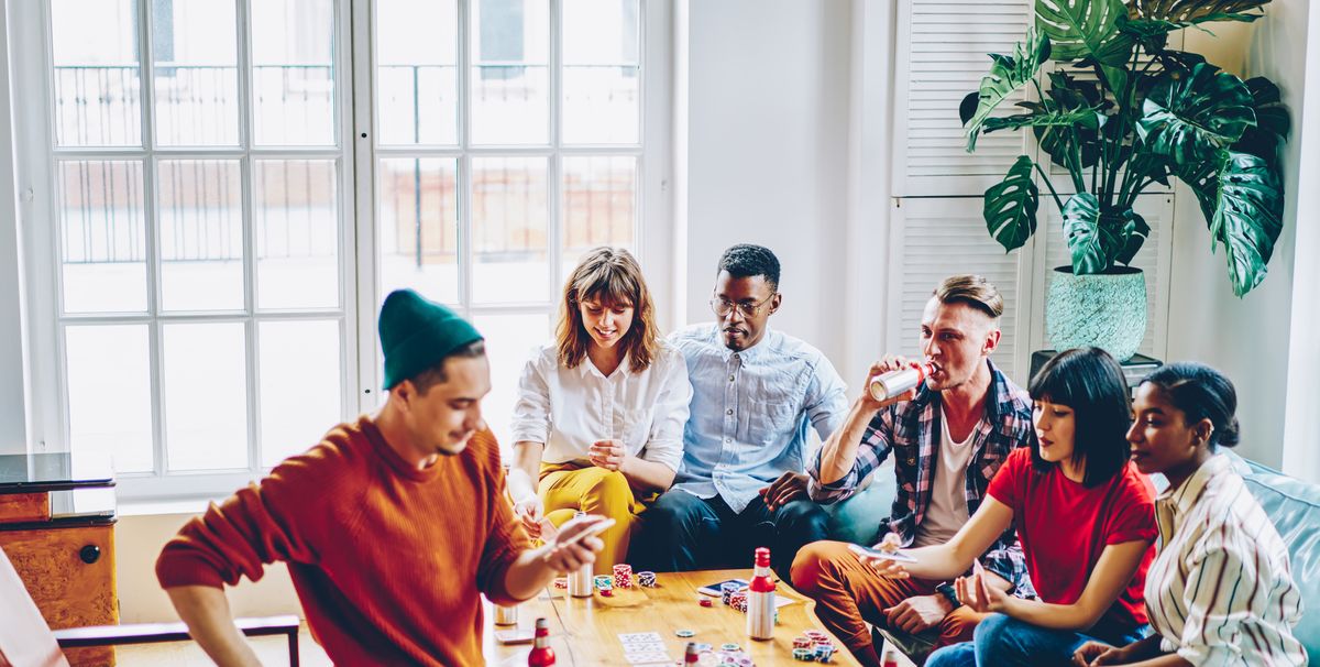 Diverse cheerful hipster guys drinking beverage and playing cards at table with chips for poker in modern apartment.Positive casual dressed multicultural friends spending free time together in flat