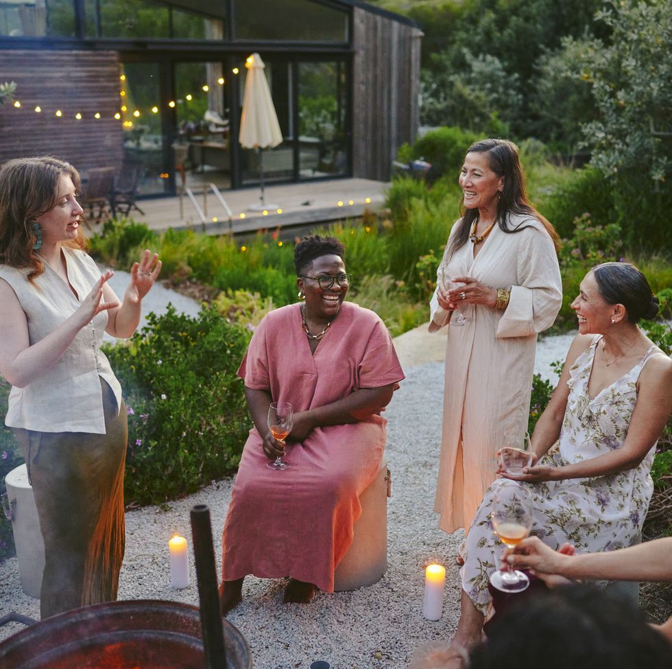 diverse women laughing over drinks around a fire pit during a garden party