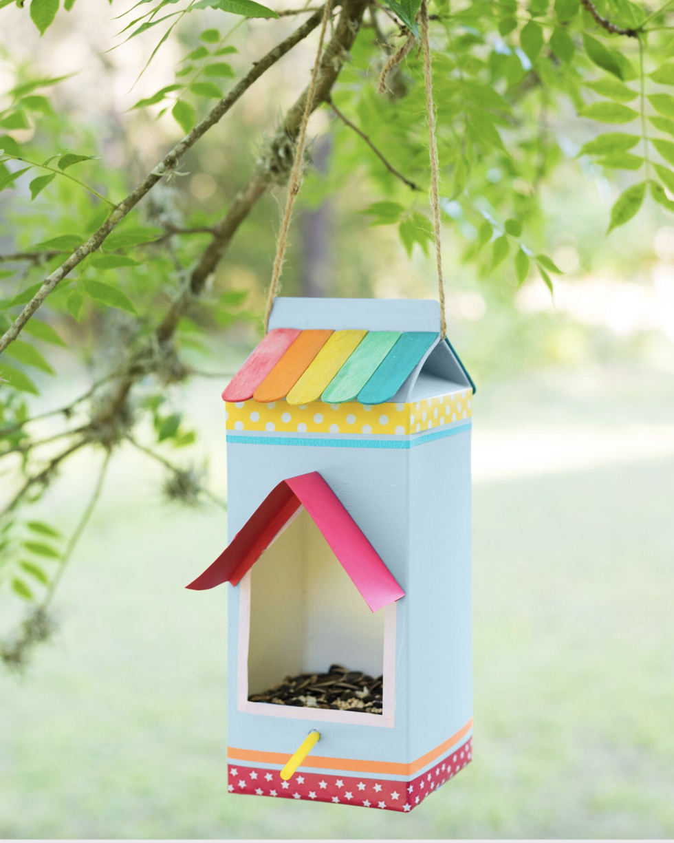 a milk carton decorated with paper washi tape and colorful popsicle sticks turned into a bird feeder hanging from a branch