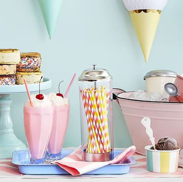birthday decoration ideas for an ice cream social themed party at home, including a paper ice cream cone bunting above a dessert table, ice cream sandwiches displayed on an aqua cake stand, milk shakes with pink and yellow striped paper straws, pints of ice cream in a pink enamelware tub of ice, and a glass canister of pink and white candy coated pretzels