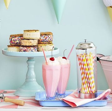 birthday decoration ideas for an ice cream social themed party at home, including a paper ice cream cone bunting above a dessert table, ice cream sandwiches displayed on an aqua cake stand, milk shakes with pink and yellow striped paper straws, pints of ice cream in a pink enamelware tub of ice, and a glass canister of pink and white candy coated pretzels
