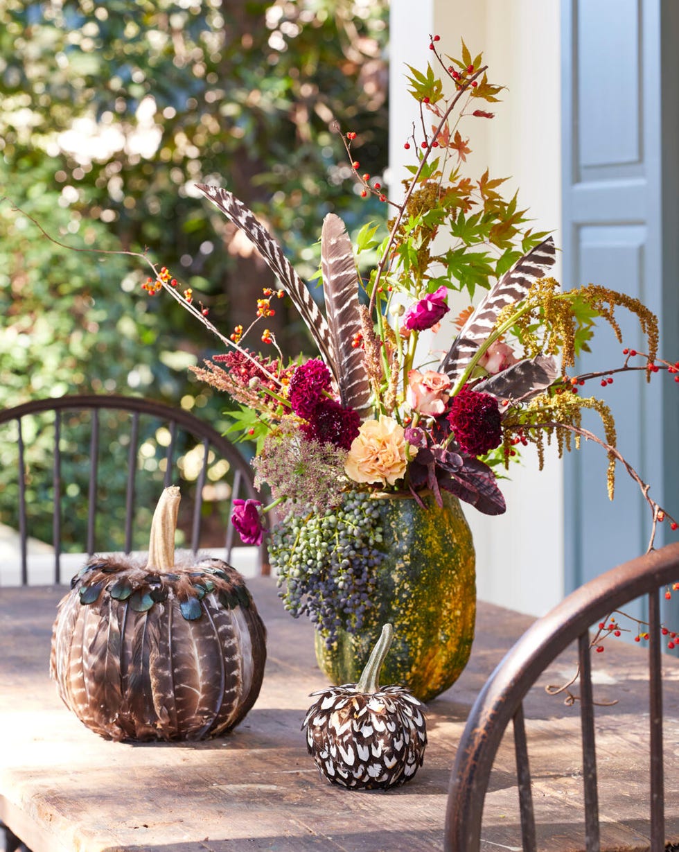 a tall pumpkin filled w ith flowers and feathers on a table with two smaller pumpkins that are completely covered in feathers