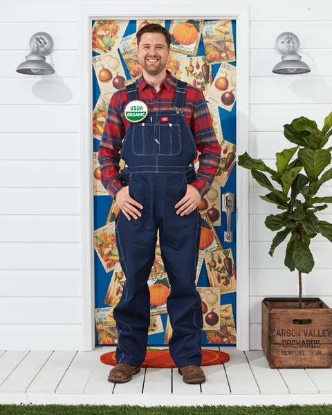 man wearing farmers costume with overalls, plaid shirt, usda organic button standing in front of door decorated for halloween
