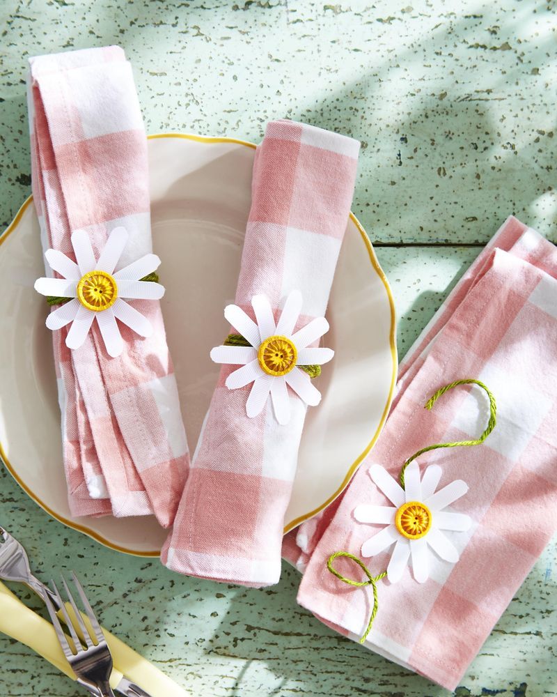 Homemade daisy napkin rings made of white ribbon, yellow buttons, and green twine tied to a pink and white gingham napkin