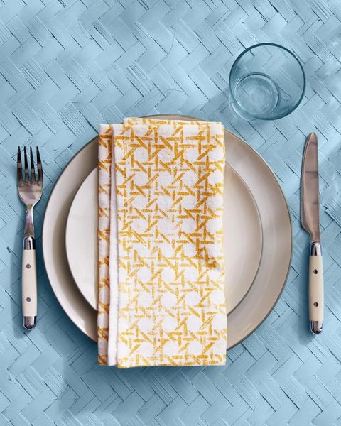 white napkin stamped with a yellow cane pattern folded on top of a simple place setting on a pale blue woven surface