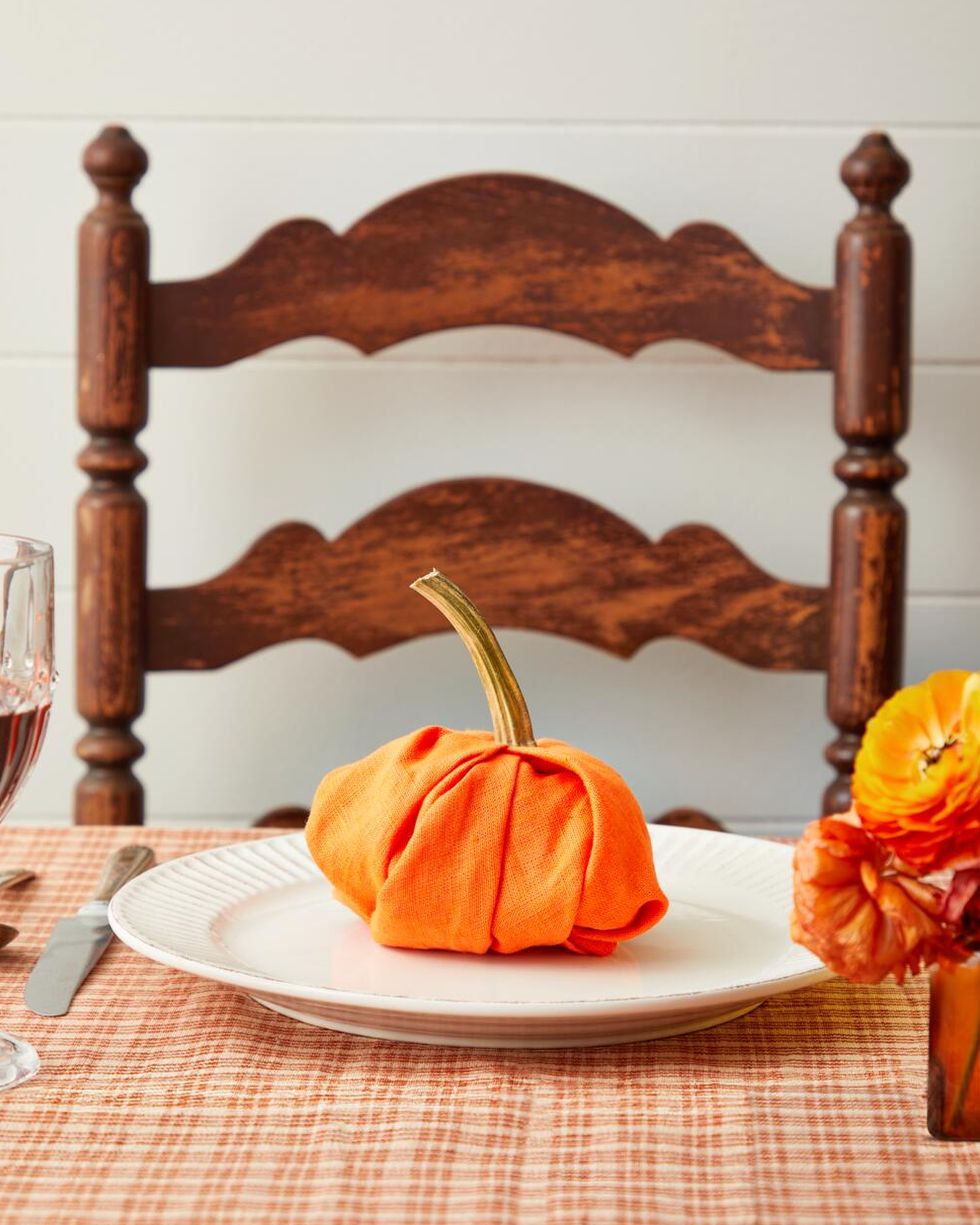 orange linen napkin folded into the shape of a pumpking iwth a real pumpkin stem on top set on a plate on a set table