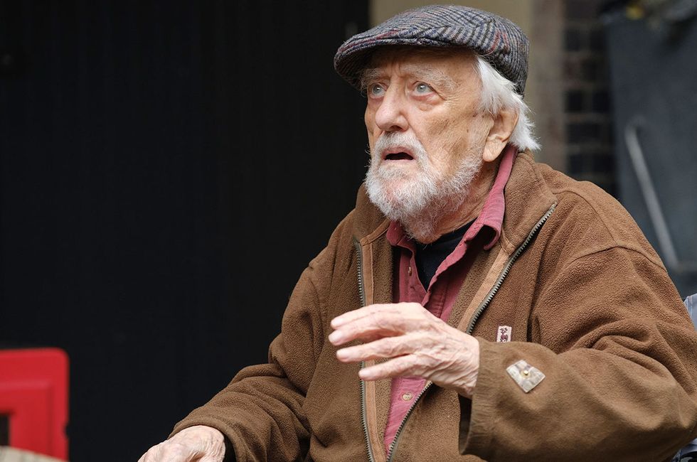 Bernard Cribbins en Doctor Who, un hombre mayor sentado en una silla de ruedas mirando a alguien fuera de cámara, tiene cabello y barba grises y usa una gorra plana con un abrigo marrón.