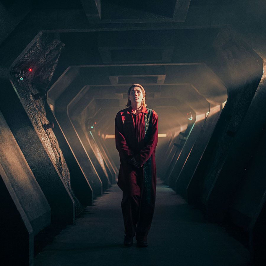 jodie whittaker in an all red outfit walks down a long foreboding corridor in a promo shot for doctor who revolution of the daleks