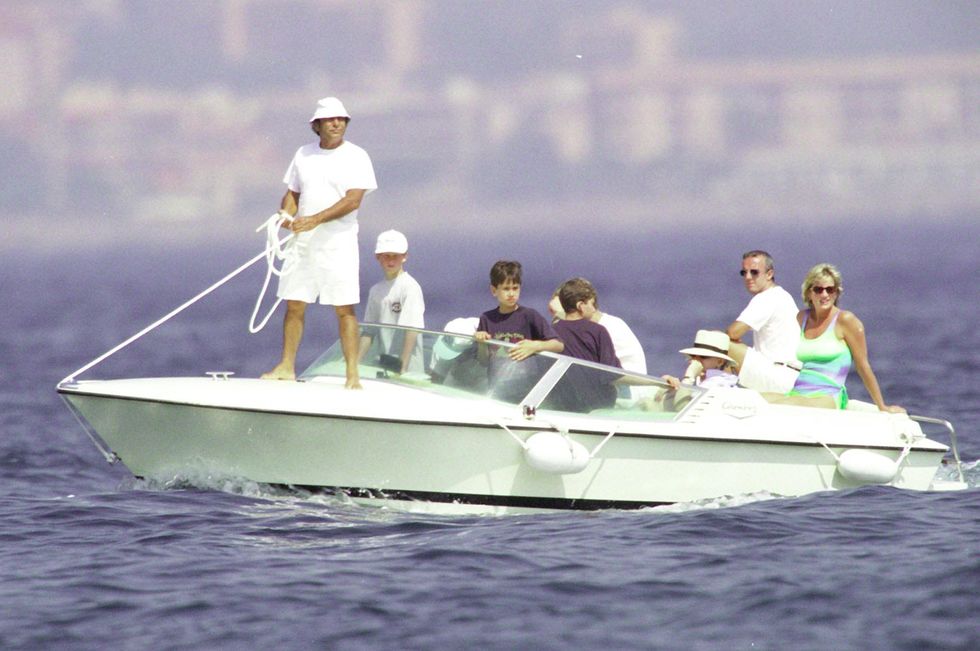 princess diana sits at the back of a boat in a green and blue swimsuit, several other people and children are onboard including a man holding a rope on the front of the boat